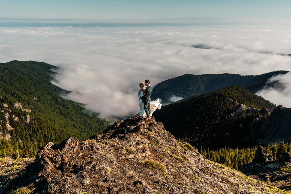 a couple in their wedding attire embrace at the top pf a mountain. they are out on the edge of a cliff and a cloud inversion swirls beneath them.