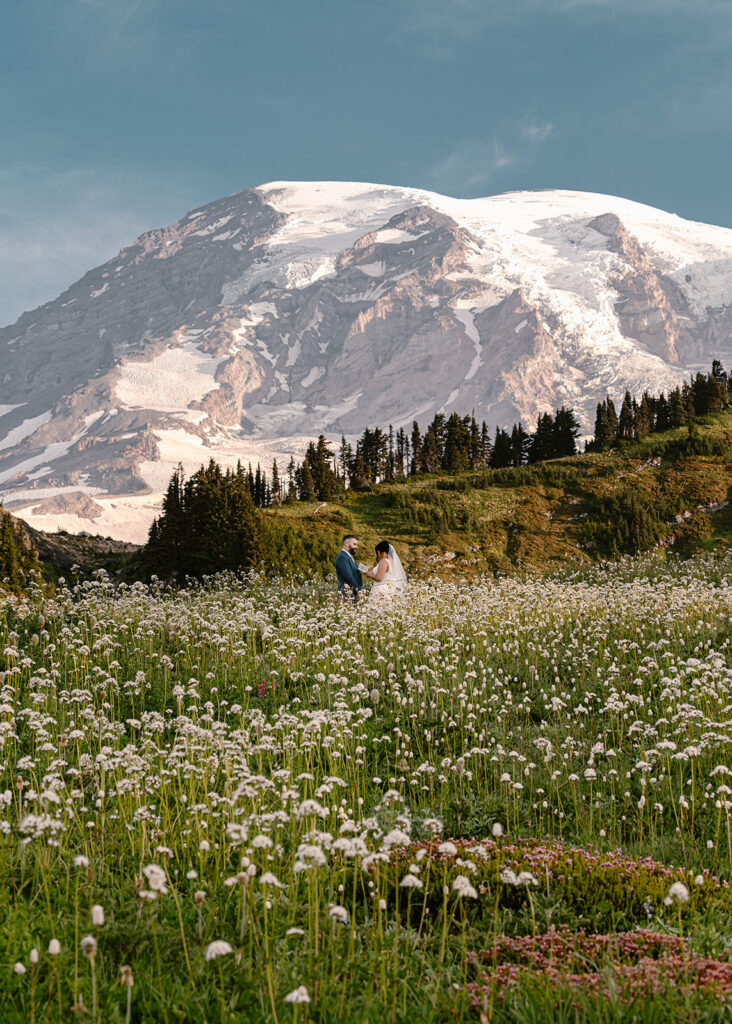 a couple stands in a field of wildflowers. The flowers fill the bottom third of the frame and rainier fills the top. The couple is in their wedding attire as they read vows to each other