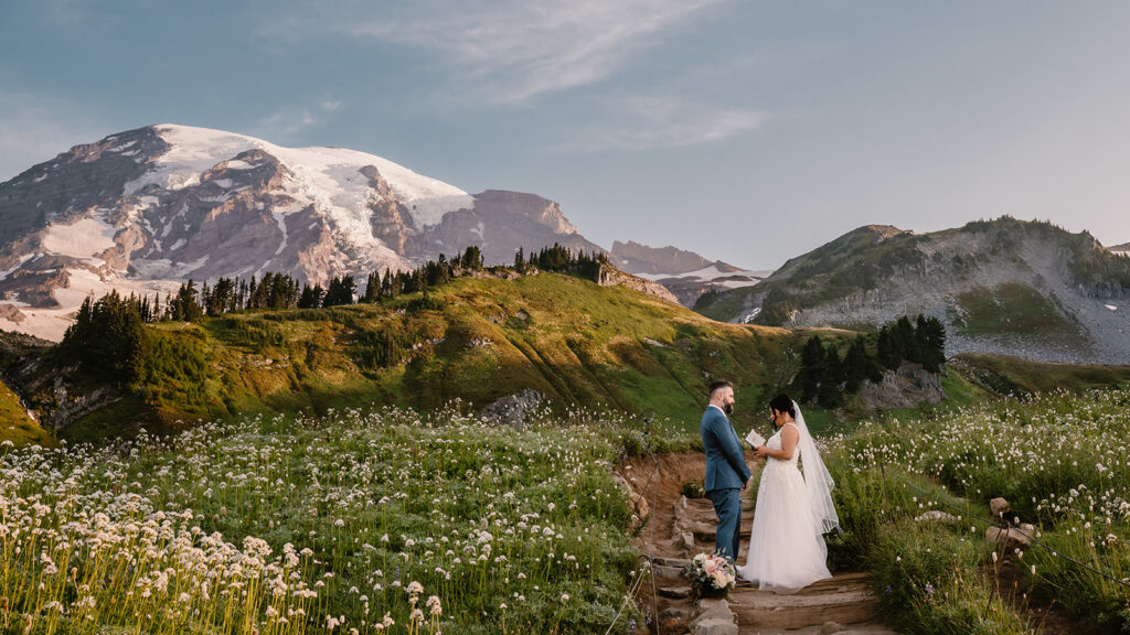 a bride reads her vows to her groom on a dirt path. in the upper left frame mount rainier rises. on either side of the trail, they are surrounded by wildflowers