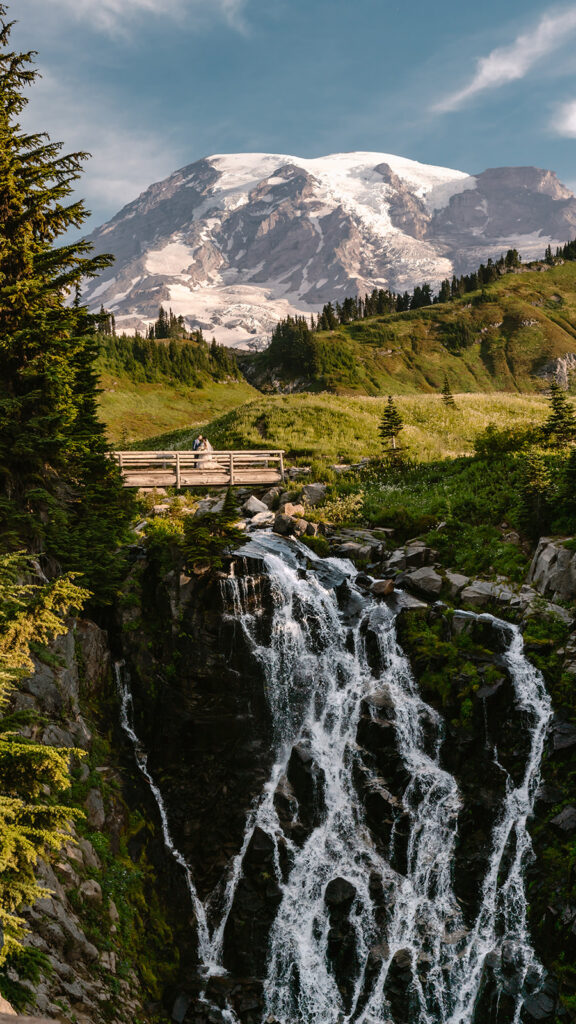 a long vertical shot of a bride and groom embracing on a wooden bridge. Mount rainier rises behind them and Myrtle Falls gushes beneath them