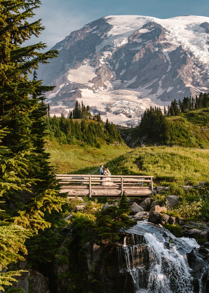 a couple in their wedding attire stand on a stunning wooden bridge. Mount Rainier rises in the background and a waterfall cascades under them