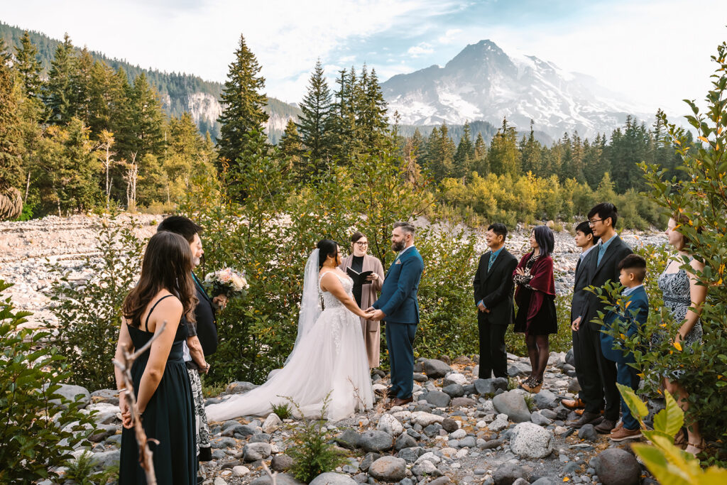 a wide shot of a mount rainier micro wedding ceremony. A couple in wedding attire is surrounded by their guests a long a rocky river bed. we see a stunning rainier in the background
