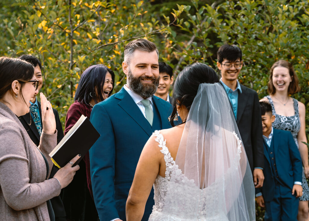 a groom gazes lovingly at his bride during their mount rainier micro wedding ceremony. In the background we see their guests smiling