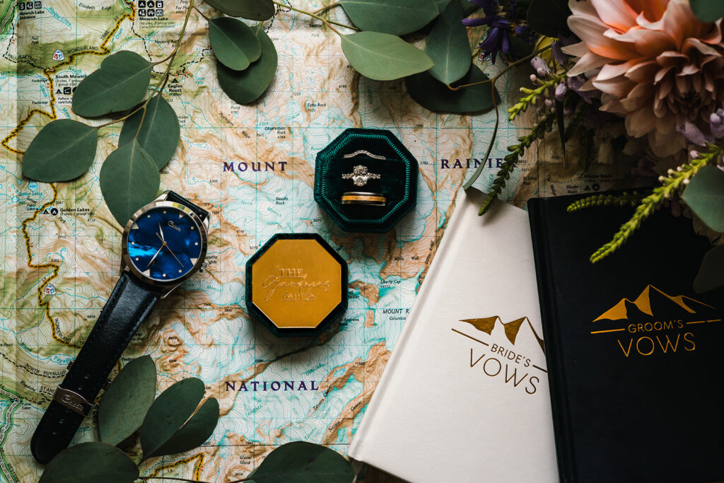 a detail shot of a national geographic map of mount rainier. on each corner we see the halves of a ring box. the box is emerald green and we see the wedding bands sparkling. A watch rests on the map beside black and white vow books