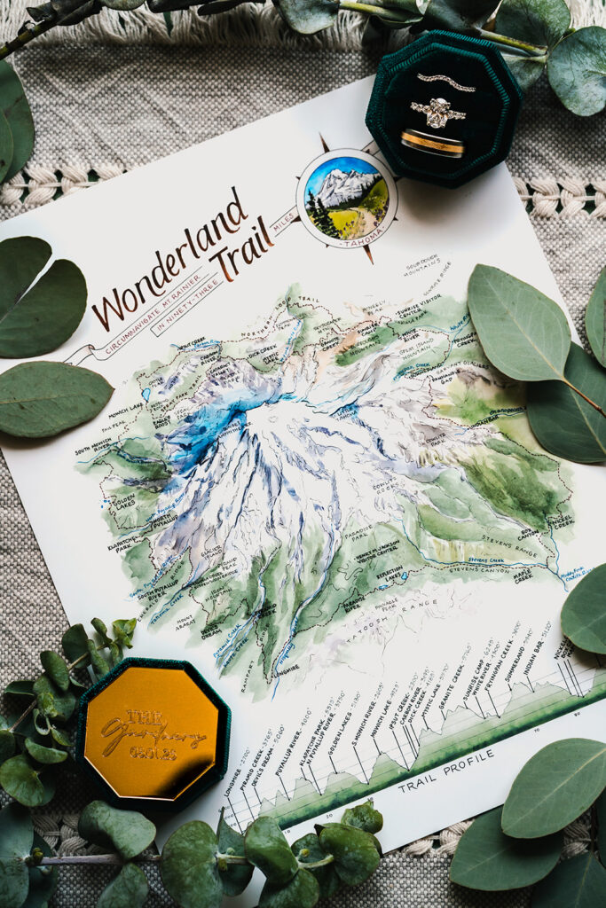 a detail shot a hand drawn map of mount rainier. on each corner we see the halves of a ring box. the box is emerald green and we see the wedding bands sparkling