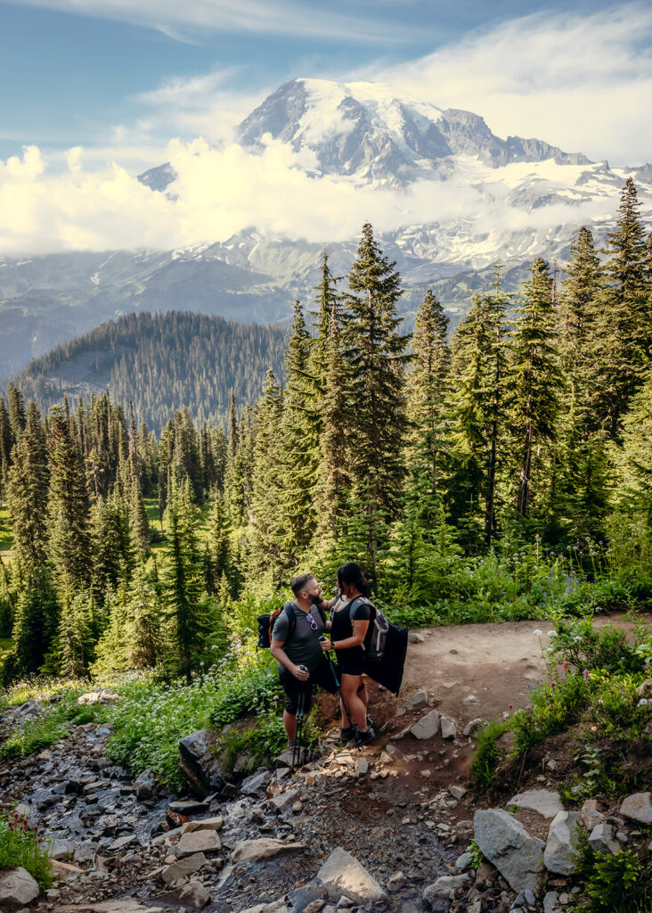 a couple in hiking attire rests and gaze lovingly at each other in the shade a long a rocky trail. Mount Rainier stands behind them