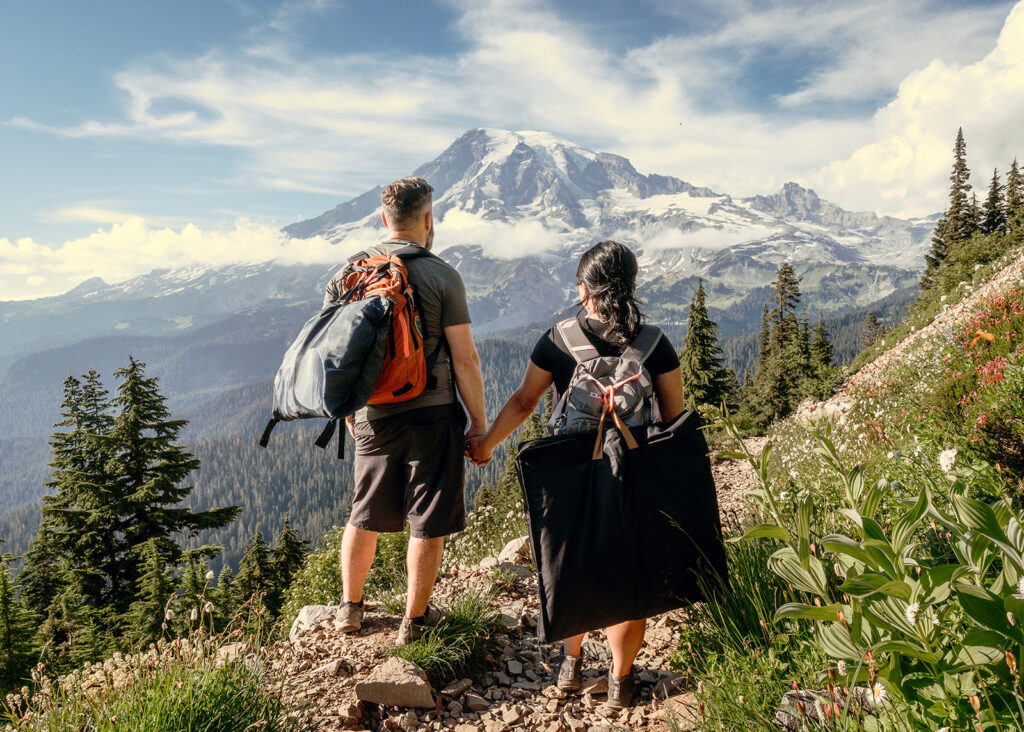 a couple hikes with their wedding attire attached to their packs. they hold hands as they gaze out at mount rainier