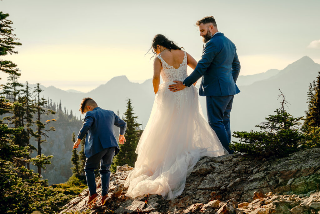 a bride and groom watch as their son begins to explore the rocky trail. A hazy scene of mountain ridges spreads in front of them
