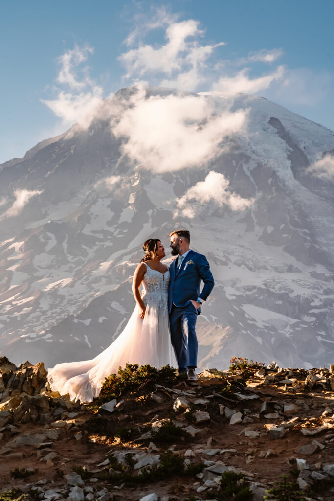 a couple in their wedding attire stand in front of a massive mount rainier gazing lovingly at each other