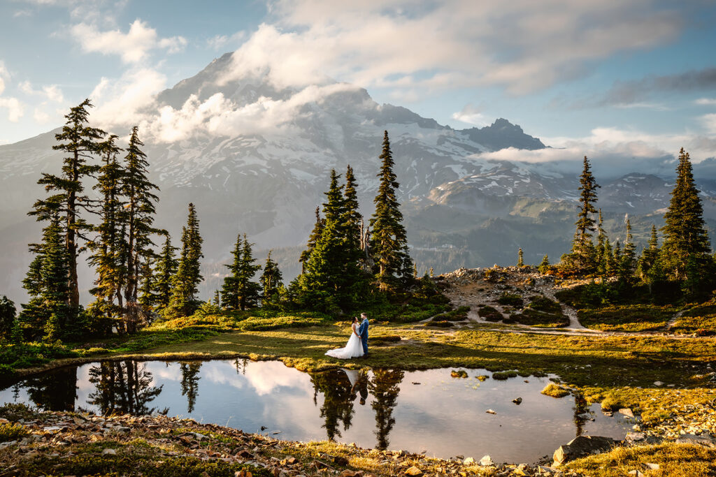 a couple stands near a reflective tarn in their wedding attire. a soft mount rainier rises behind them painted with pearly clouds and soft sunlight. the scene is also reflected below them in the tarn
