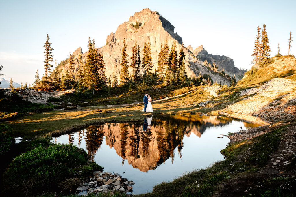 a couple stands near a reflective tarn in their wedding attire. a golden pinnacle peak rises behind them painted with golden sunlight. It is also reflected below them in the tarn