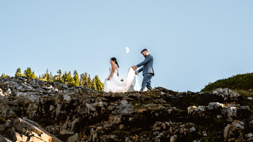 a bride explores and her groom carries the train of her dress. It is spread out as they walk, a short distance between them. A perfect half moon rises above them, perfectly centered between them.