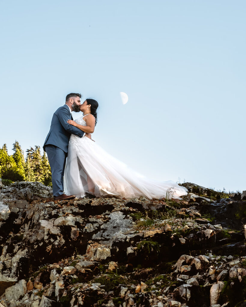 a bride and groom kiss deeply. They are on rocky terrain, and there is a perfect half moon directly above them
