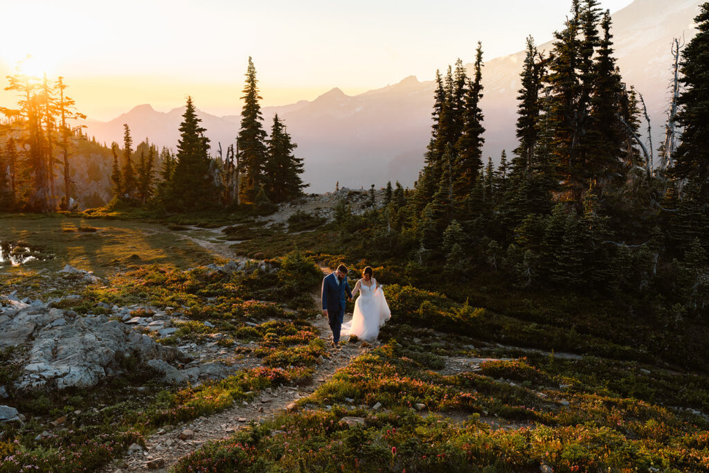 a bride and groom explore a trail in their wedding attire. the sun sets behind them creating glowing pink, purple, and golden hues
