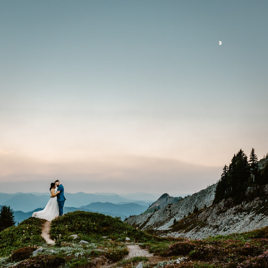 a bride and groom share a kiss in a blue and green landscape as blue hour sets in. in the top third of the frame is a tiny, pearly half moon