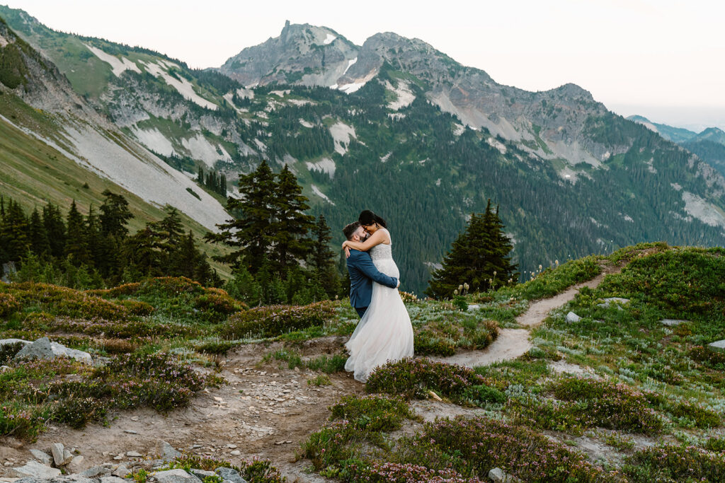 a groom lifts his bride as the sun begins to set. mountains rise behind them and the trail spreads and winds beneath them