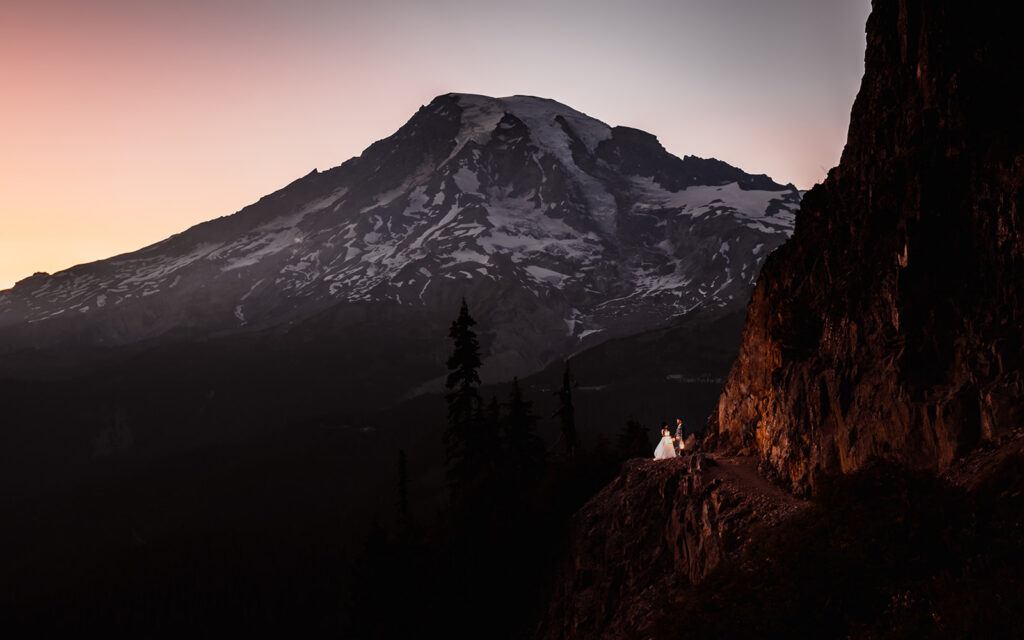 a landscape shot of a bride and groom kiss under the view of rainier. It is dusk now and the only light that remains is the subtle blue glow of the mountain and the warm light of their lanters
