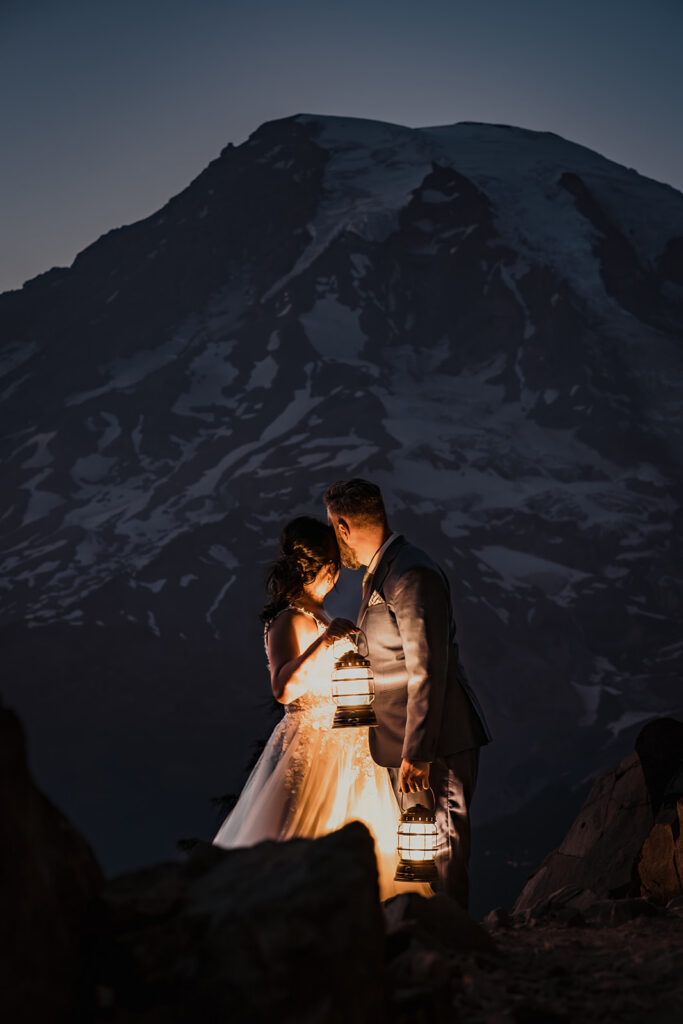 at the end of their mount rainier micro wedding, a couple stands cheek to cheek in their wedding attire. They hold lanterns as they gaze out at a stunning mount rainier painted in purple light