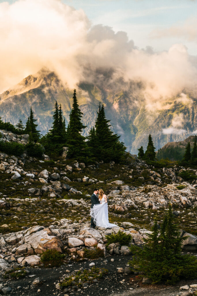 a bride and groom dance forehead to forehead perfectly aligned under the highest point of a mountain. Pearly, golden clouds billow above them