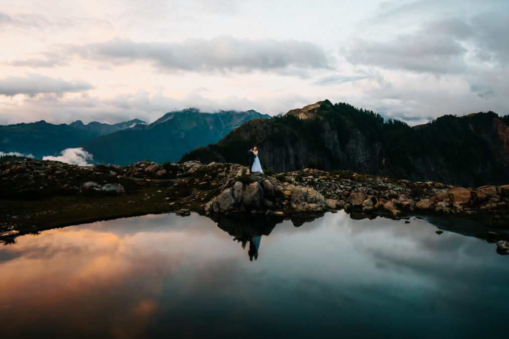 a stunning shot of a bride and groom standing above a reflective tarn. We see the stunning sunset reflected back with its deep blues