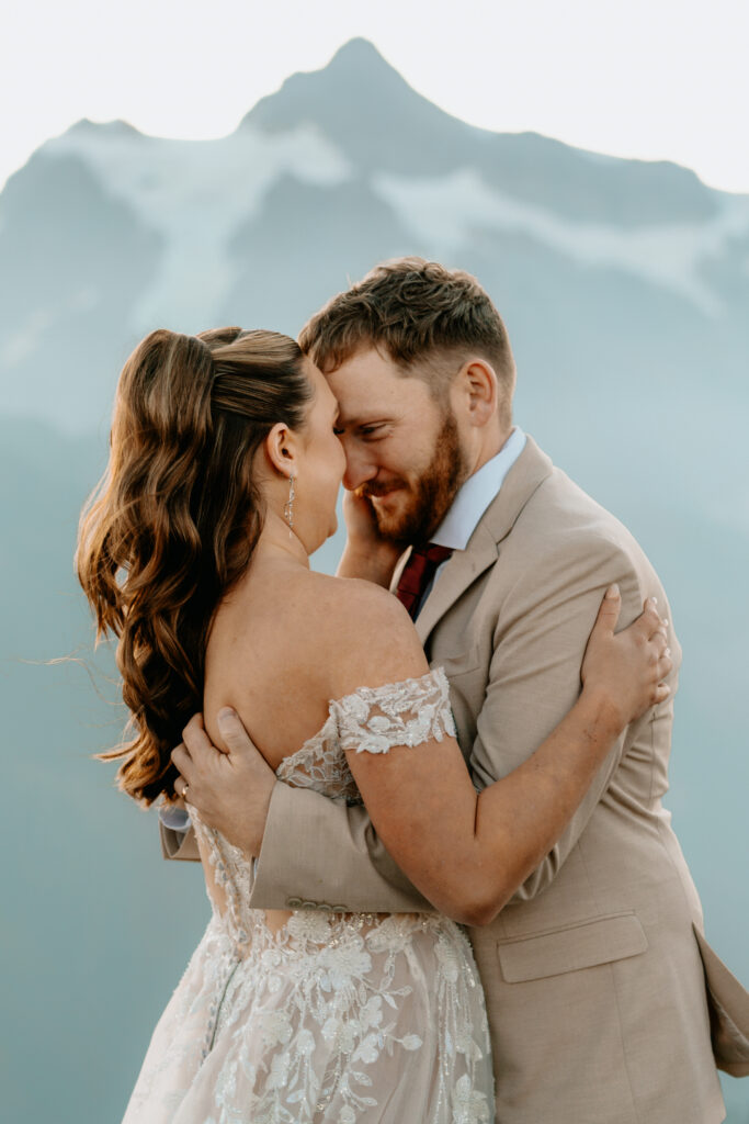 a groom pulls his bride in close and they smile sweetly at each other. we see the pale blue face of a mountain behind them and layers of snow in the ridges