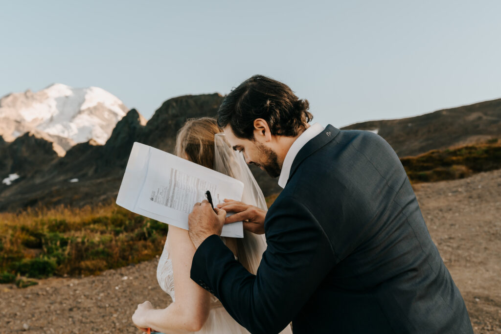 a groom signs his marriage license. They are in the backcountry and a glacier can be seen in the background. His bride turns around and he places the washington marriage license on her back to sign