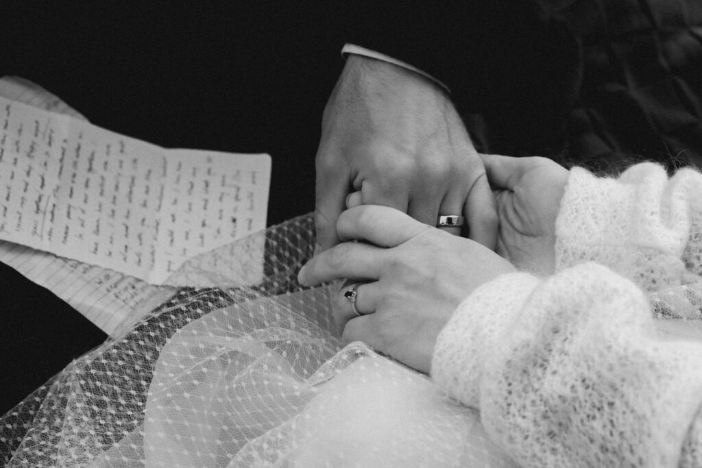 a black and white image of a couple in their wedding attire holding hands with their new wedding bands. Blurry vows, written on notebook paper can be seen in the corner of the frame