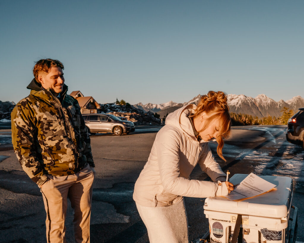 a bride and groom are bundled in coats in a snowy parking lot. The wind whips their hair around as the bride signs her marriage license on top of a yeti cooler. Her groom smiles broadly at her and mountains rise up jagged behind them