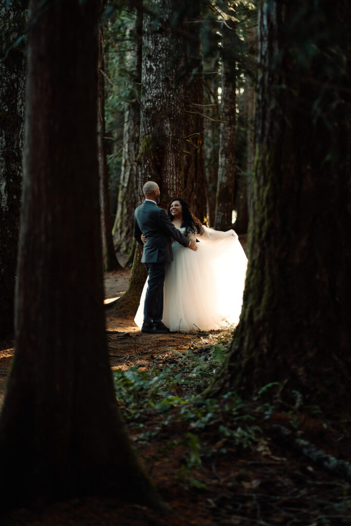 a couple dances in the forest. Light streams gently through the forest canopy and highlights the fabric of the brides dress as she gazes lovingly at her husband
