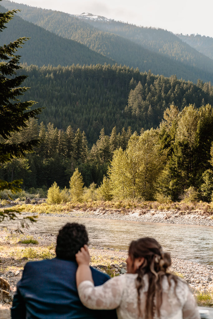 a bride caresses her grooms cheek as they gaze out at the river a head of them. They are blurred and the focus is on the river that boarders their stay at Mount Rainier