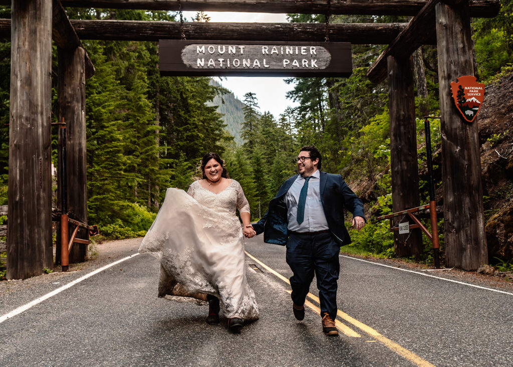 a couple in their wedding attire run towards the camera smiling widely. Above them is the entrance sign to Mount Rainier National Park. 