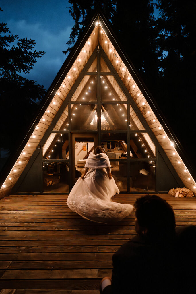 a bride twirls under the peak of their stay near Mount Rainier. She is illuminated by the patio lights that follow the shape of the cabin. in the bottom right frame, we see the silhouette of her groom watching her dance  