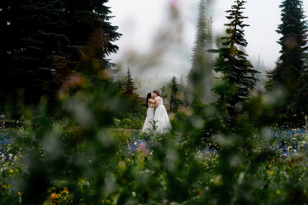 Two brides, in their wedding dresses, kiss deeply in a sea of wildflowers at Mount Rainier. They are framed between the dotted colors of the flowers. The skies are gray, the atmosphere is moody and ethereal. 