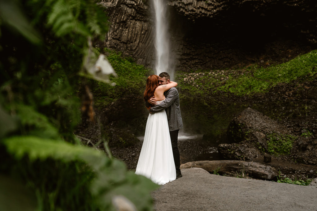 a couple embraces during their waterfall elopement surrounded by greenery with gushing falls in the background