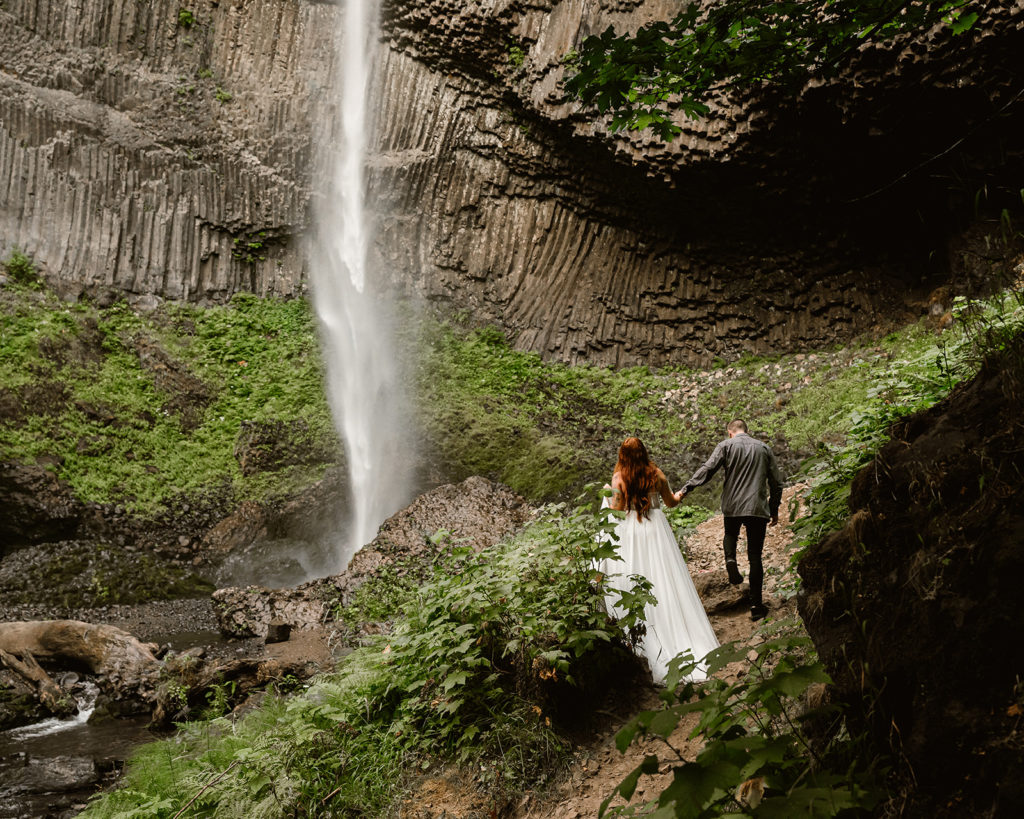 Couple in wedding attire walks through forest towards a waterfall during their waterfall elopement.