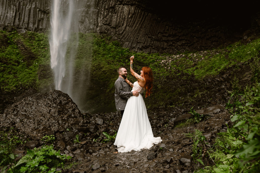 Couple in wedding attire cheers in front of waterfall during their waterfall elopement.