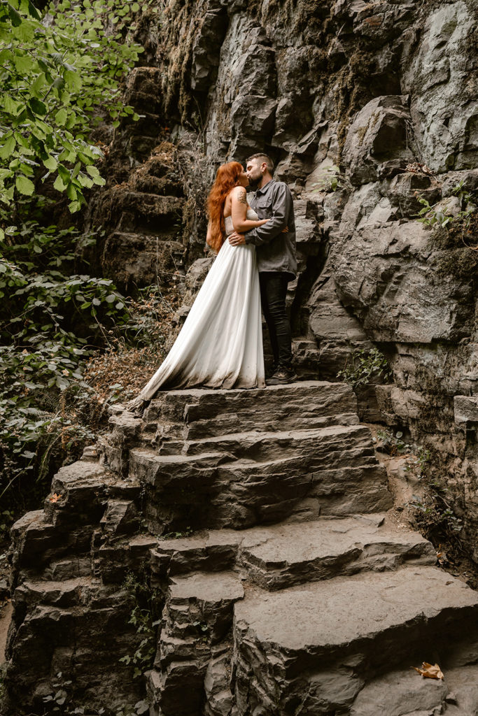 Couple kisses in wedding attire standing on basalt rock formations during their waterfall elopement.