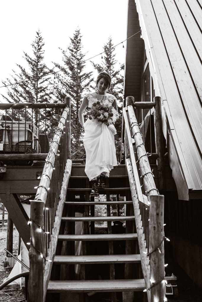 black and white photo of a bride walking down the steps of their aframe cabin holding her boquet during her mt. rainier wedding  
