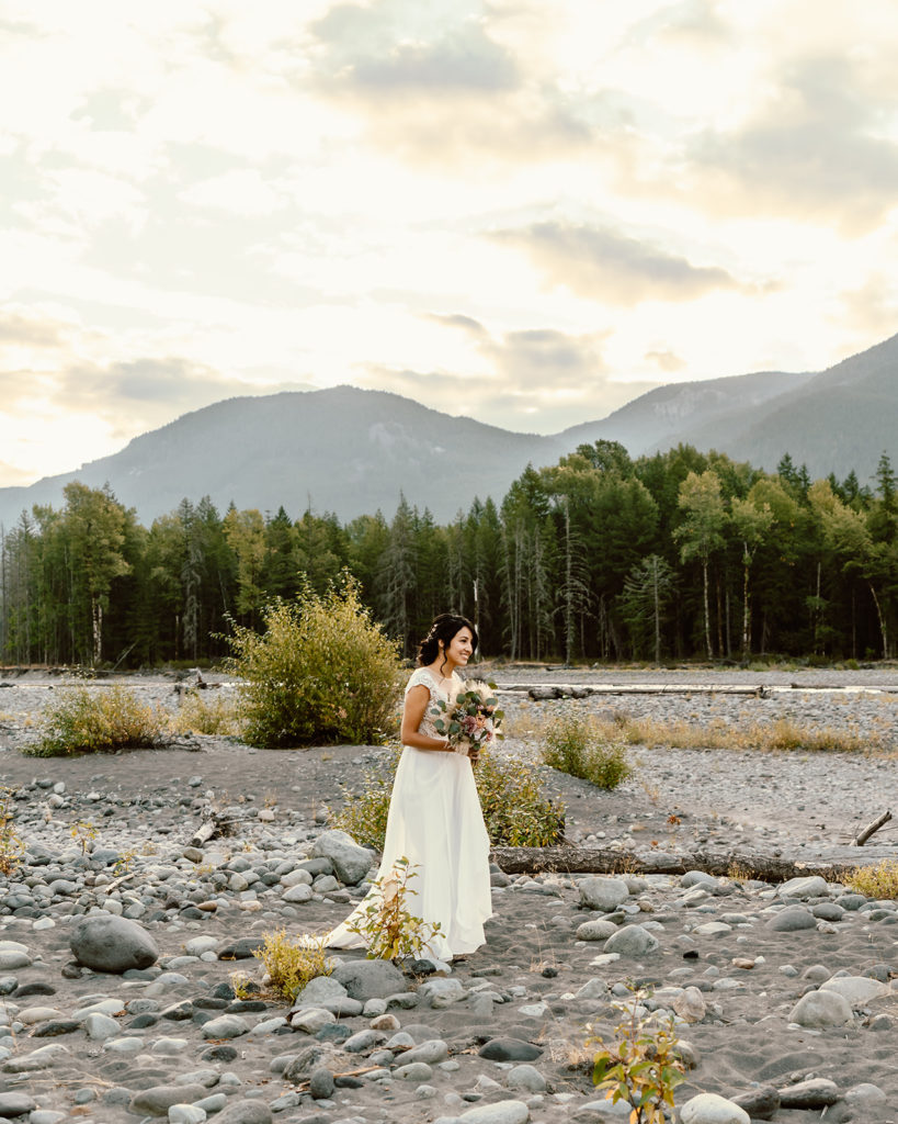 Bride walks along river bed at sunrise with mountains and trees in the background during her Mt. Rainier wedding 
