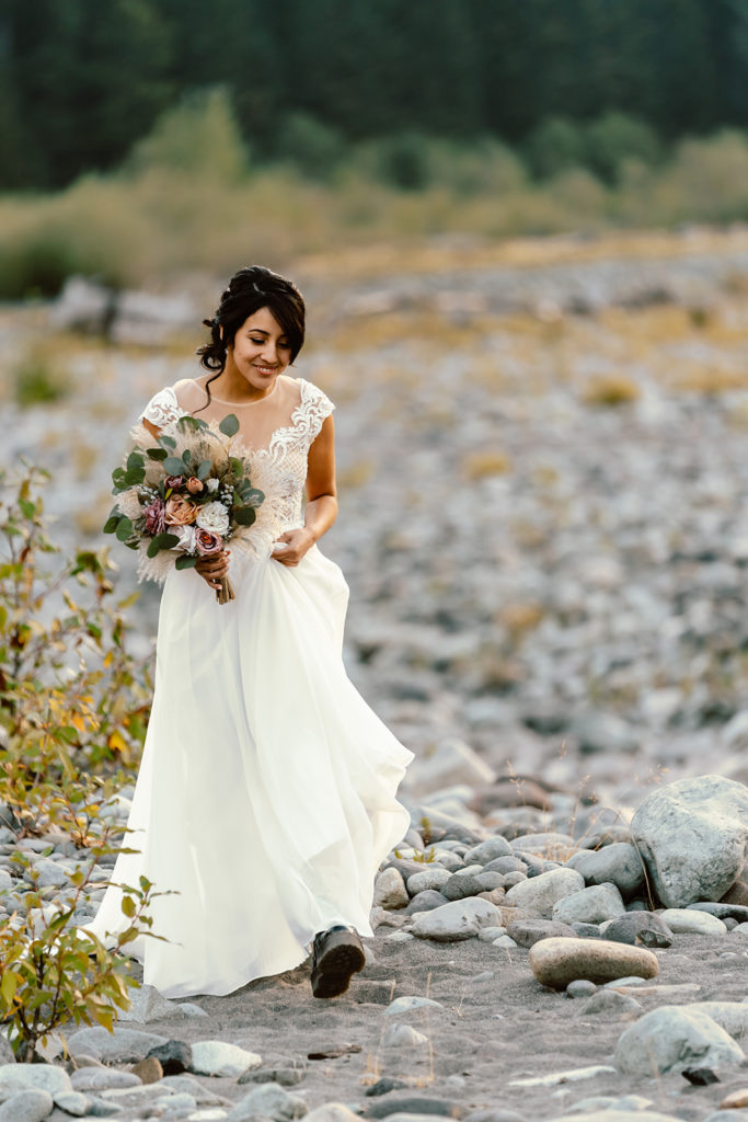 Bride walks along river bed at sunrise with mountains and trees in the background during her Mt. Rainier wedding 