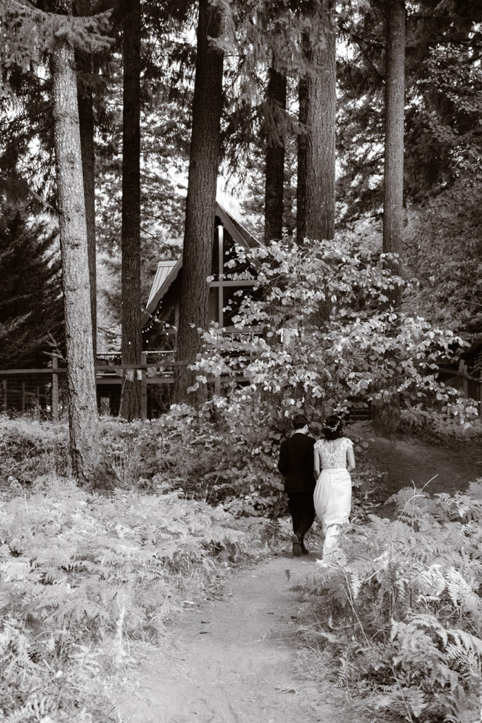 black and white photo of bride and groom walking towards their A-frame cabin nestled in the woods during their mt. rainier wedding