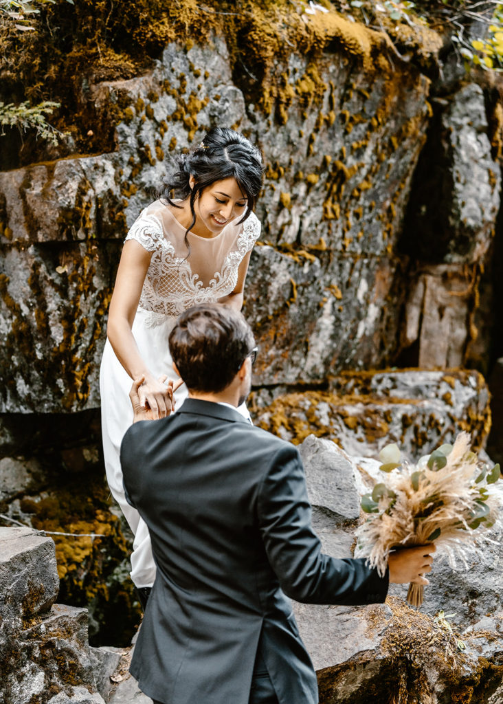 Groom holds bouquet as he helps his bride climb down to their mt. rainier wedding location 