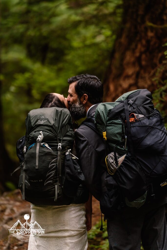 A bride and groom hike in their wedding attire on a forested trail wearing their packs. They kiss during their Washington Elopement.