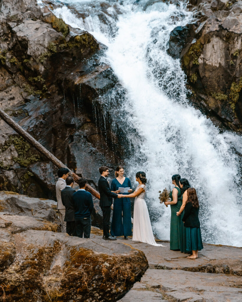 After planning their elopement timeline, a couple and their guests  have a ceremony on a mossy cliff with a raging waterfall behind them.