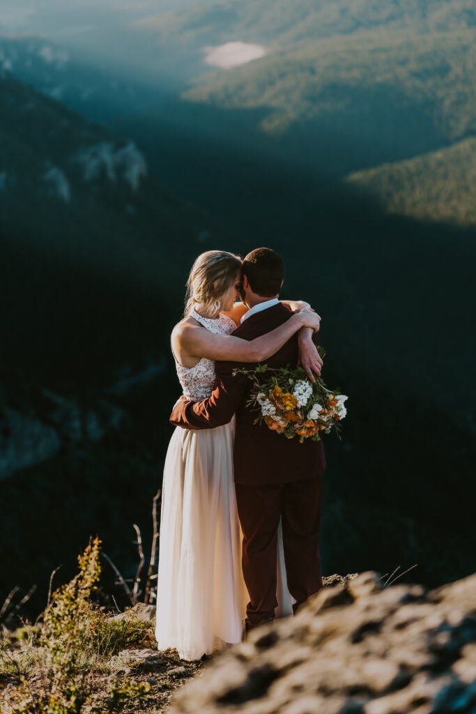 After considering elopement vs wedding, a couple in wedding attire, embrace at sunrise after exchanging vows on a mountain top. A bride holds a bright orange bouquet that pops against the grooms deep maroon suit. 