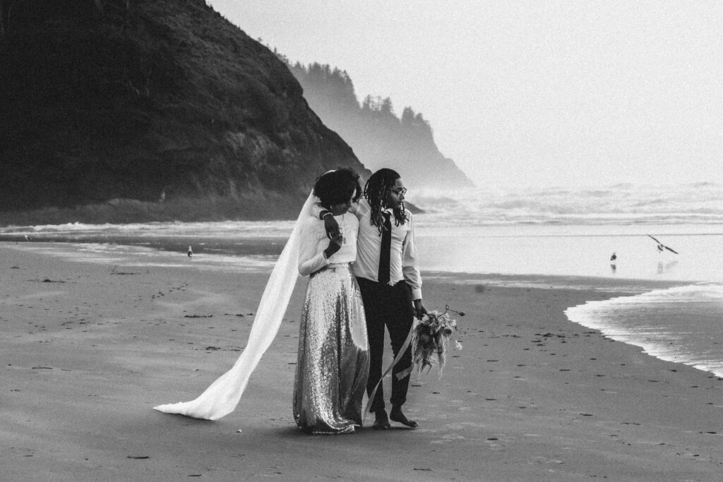 After considering elopement vs wedding, a couple in wedding attire explores the foggy, rocky coastline. seagulls fly beside them, the reflection of the tide curves before them, and a large sea-rock with coastal trees serves as the background to their elopement day.  