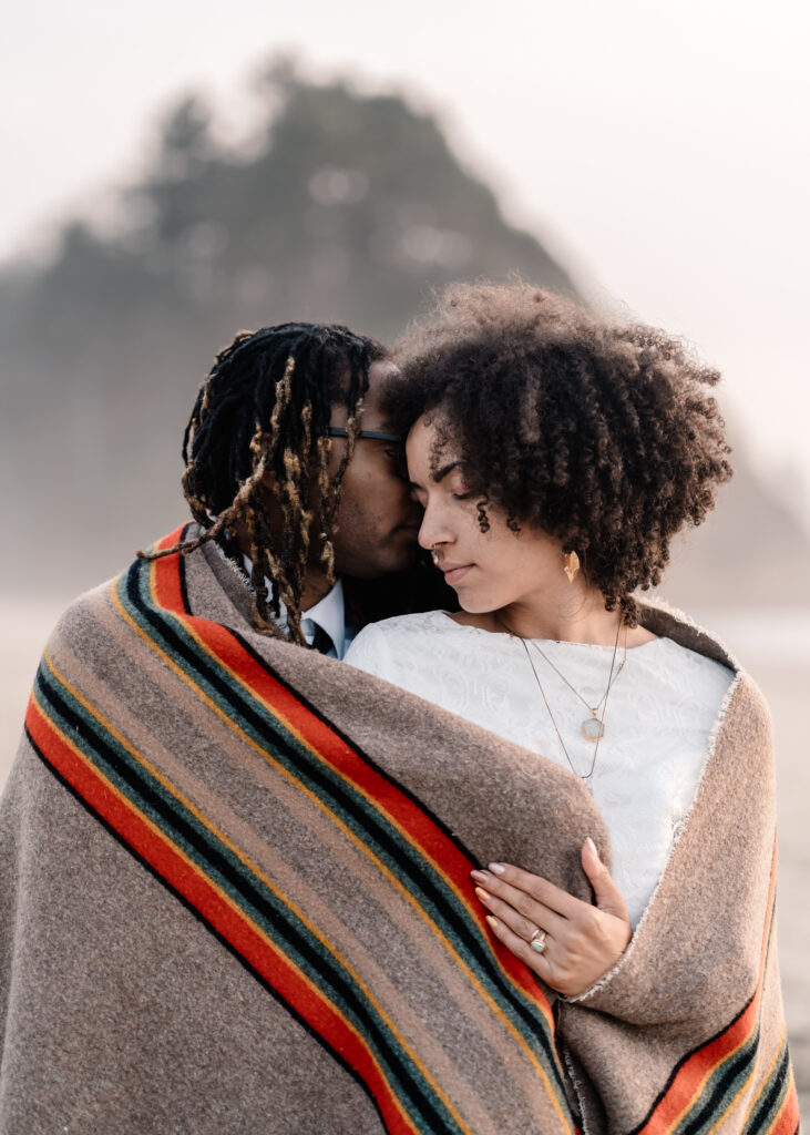 After considering elopement vs wedding, a couple in wedding attire snuggles up in a striped Pendleton camping blanket, nuzzling cheeks during their elopement in front of a large haystack rock on the coast.