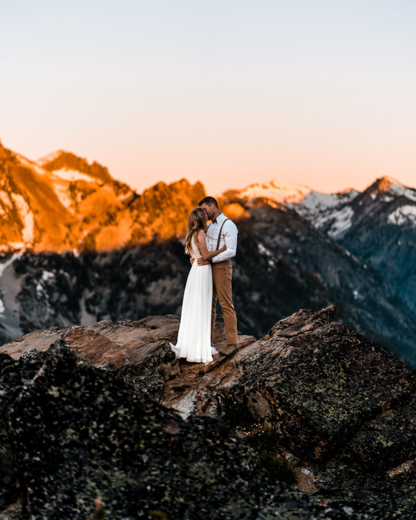 During their elopement timeline, a couple in wedding attire, embraces on top of a rock cliff surrounded by a ridge-line highlighted with blue shadows and orange alpenglow.