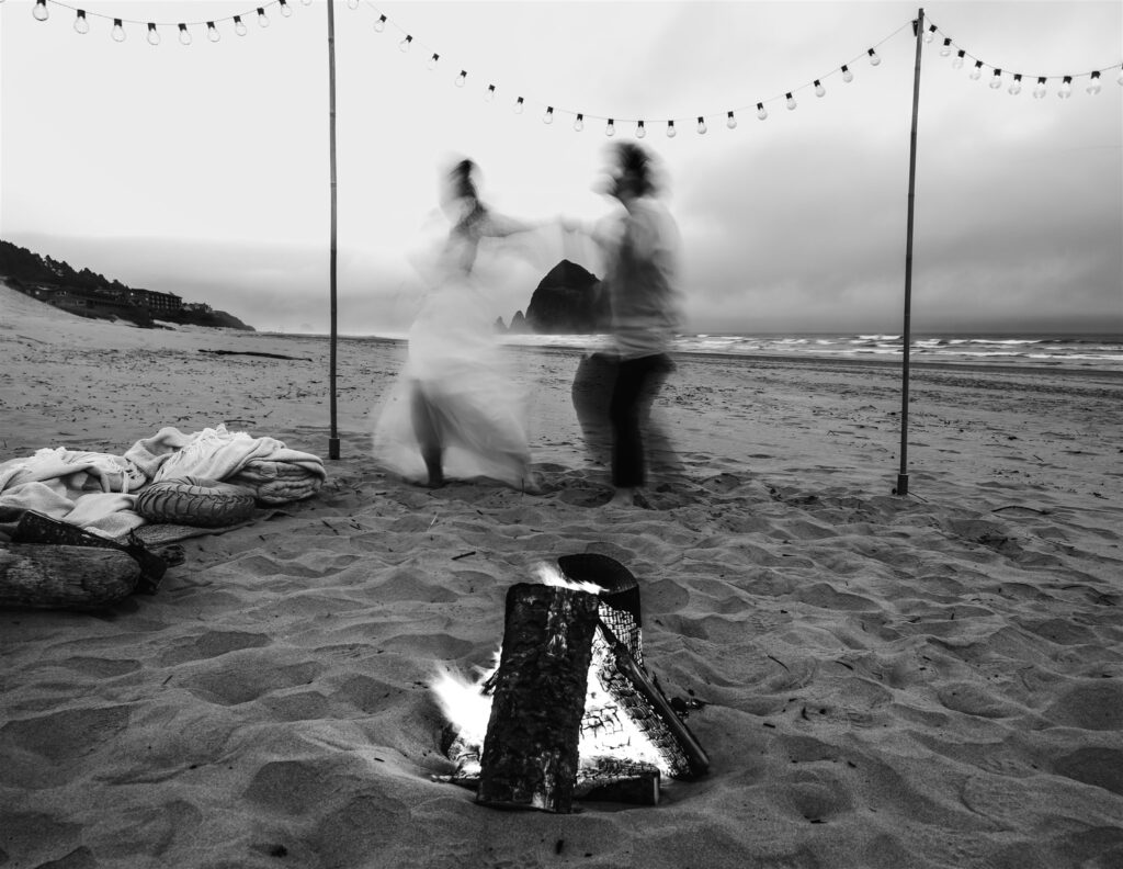 After considering elopement vs wedding,  a couple is blurry with movement from their dance party in front of a crisp silhouette of a giant haystack rock on Cannon Beach. They dance surrounded by a cozy bonfire setup surrounded by cozy blankets, pillows, and string lights.  