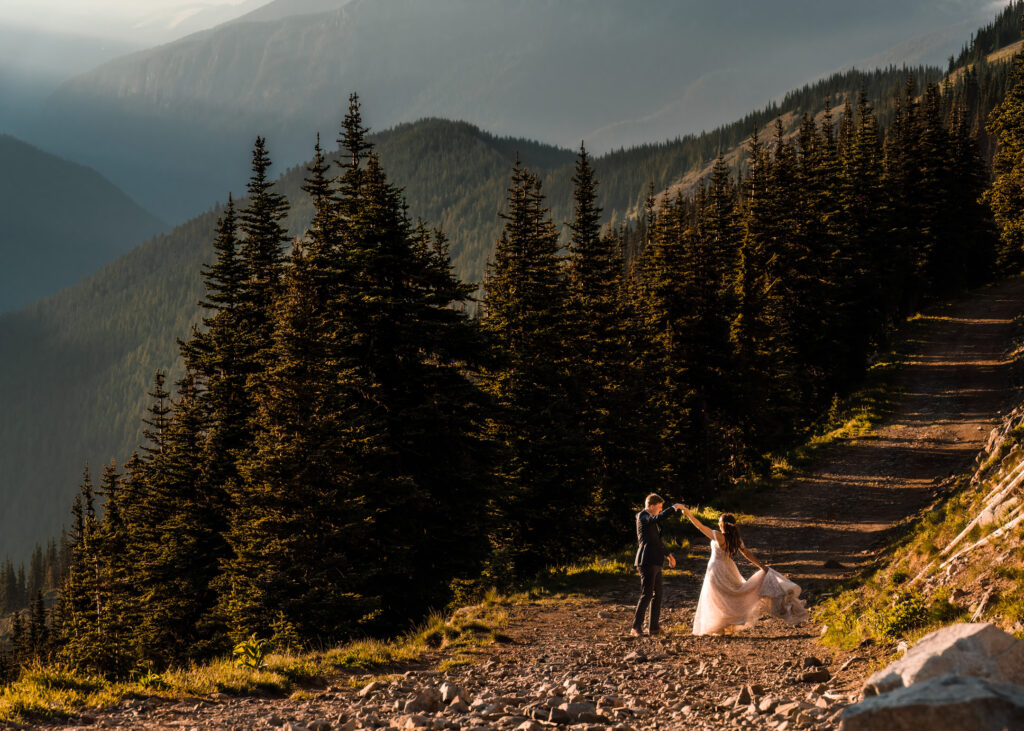 After considering elopement vs wedding, dances on a wide gravel path above a sunlit mountain range, surrounded by massive pines and a blue-green mountain range